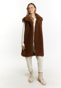 Brun, fyldig ærmeløs frakke med krave; båret over en lys top og bukser. Beige snørresko fuldender outfittet.