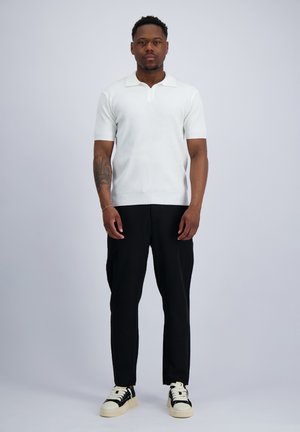 Ein Mann steht nach vorne gerichtet, trägt ein weißes Kurzarmhemd, schwarze Hose und schwarz-weiße Turnschuhe vor einem schlichten, hellen Hintergrund.