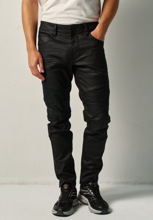Hombre con pantalones negros texturizados y zapatillas negras, de pie con una mano en el bolsillo frente a un fondo liso.