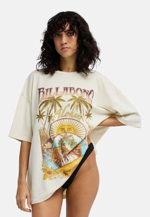 Kobieta z kręconymi włosami ubrana w białą, oversize koszulkę Billabong z tropikalnym nadrukiem i czarne majtki bikini, ręka na biodrze.