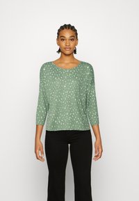 Top verde de mangas três quartos com um padrão de pontos brancos espalhados, decote redondo e uma textura suave, combinado com calças pretas.