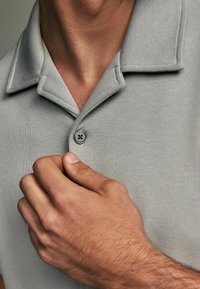Polo shirt grigio chiaro realizzato in tessuto strutturato, con colletto a coste, patta con due bottoni e un motivo sottile. Bottoni realizzati a mano vicino al colletto.