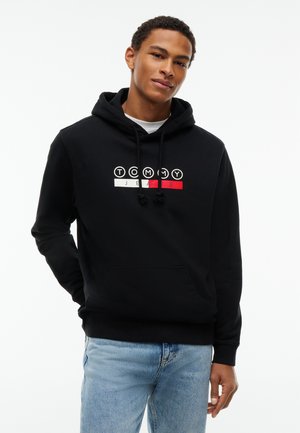 Joven con sudadera con capucha negra de Tommy Jeans y vaqueros azul claro, de pie con un fondo neutro.