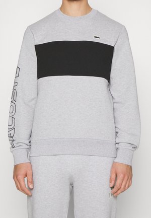 Homme portant un sweat-shirt gris clair avec une large bande noire sur la poitrine, logo Lacoste sur la poitrine gauche et texte Lacoste sur la manche droite.