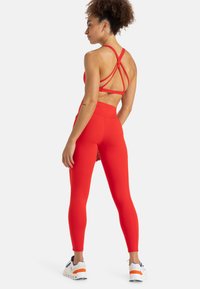 Tenue de fitness rouge comprenant un soutien-gorge de sport à dos croisé et un legging taille haute, tous deux en tissu lisse et extensible. Baskets blanches et marines.