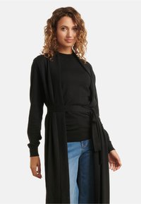 Black long cardigan with a tie waist, featuring long sleeves and an open front design. Worn over a black top and blue jeans. Smooth texture.