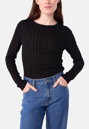 Frau trägt ein enganliegendes schwarzes, strukturiertes Langarmoberteil und hoch taillierte blaue Jeans mit Händen in den Taschen.