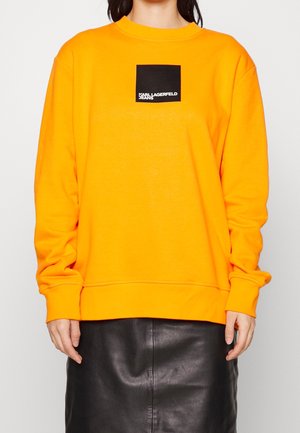 Persona vistiendo una sudadera de manga larga de color naranja brillante con un logo cuadrado negro que dice "Karl Lagerfeld Jeans", combinada con una falda de cuero negra.