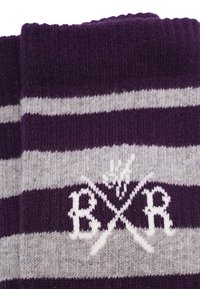 Lilla og grå stribede sokker med strikket tekstur, der har et hvidt skiltlogo med krydsede genstande og bogstaverne "B R" i midten.