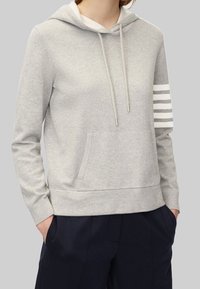 Světlá šedá mikina z jemné látky s přední kapsou, nastavitelnými šnůrkami a třemi bílými pruhy na levém rukávu.