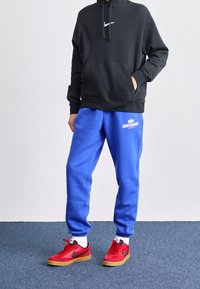 Osoba v černé mikině s bílým logem Nike, modrých sportovních kalhotách Nike, bílých ponožkách a červených teniskách Nike stojí na modrém koberci.