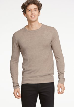 Beiger Pullover mit langen Ärmeln aus weichem Strickmaterial, mit Rundhalsausschnitt und tailliertem Schnitt, kombiniert mit dunklen Jeans.