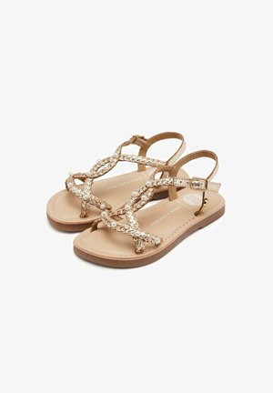 Sandalias de color dorado con correas cruzadas, adornadas con perlas blancas. Suela plana, correa ajustable en el tobillo y acabado de cuero suave.