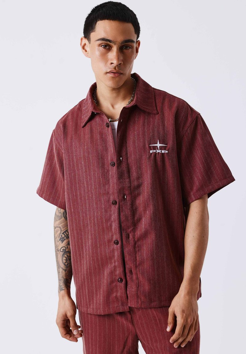 Jeune homme aux cheveux courts et foncés porte une chemise ample bordeaux à fines rayures à manches courtes et un pantalon assorti, montrant un bras gauche tatoué.