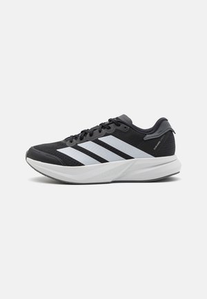 Chaussures de sport noires avec une tige en mesh respirant, trois rayures blanches et une semelle intermédiaire gris clair avec le logo "LIGHTSTRIKE".