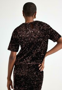 Sort top med paljetter og cut-out ryg, korte ærmer, struktureret overflade og glitrende detaljer mod en lys baggrund.