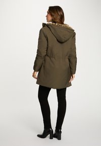 Parka verde oliva con cappuccio foderato in pelliccia, vita stretta e taglio dritto, abbinata a pantaloni skinny neri e stivali con tacco neri.