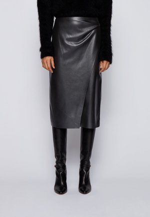 Black faux leather wrap skirt worn with black fuzzy sweater and knee-high black leather boots on plain background.