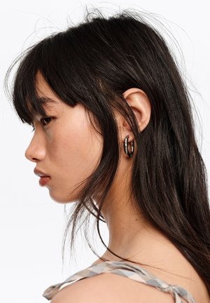 Mujer joven con el pelo largo y oscuro que lleva un pendiente de aro plateado, mirando hacia la izquierda sobre un fondo blanco.