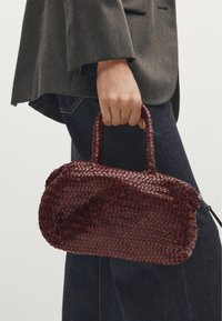 Hand holding small dark red woven leather handbag, person wearing gray blazer and dark blue jeans visible from midsection down.
