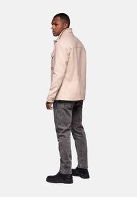Chaqueta de mezcla de lana beige con cuello y bolsillo en el pecho, combinada con jeans de mezclilla grises y botas negras, exhibida desde una perspectiva lateral.