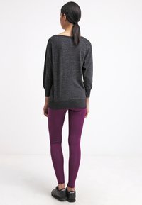 Dunkelgraues, lässig geschnittenes Langarmshirt mit rundem Ausschnitt, kombiniert mit enganliegenden, glänzenden, tiefpurpurfarbenen Leggings und schwarzen Sportschuhen.