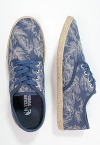 Paire de chaussures décontractées en toile bleue avec motifs de feuilles blanches, lacets bleus et semelles beiges rappelant une corde sur fond blanc.
