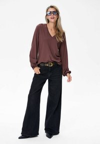 Vrouw met oversized zonnebril, bruine lange mouwen V-hals top, zwarte wijde spijkerbroek met riem en zwarte puntige schoenen, die casual staat.