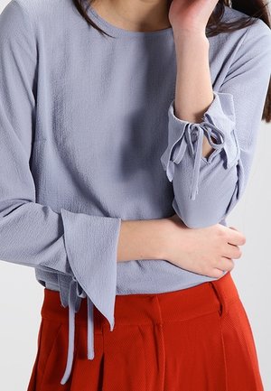 Kobieta w fakturowanej jasnofioletowej bluzce z zawiązanymi mankietami rękawów, zestawionej z wysokim stanem, plisowanymi czerwonymi spodniami. Skrzyżowane ramiona.