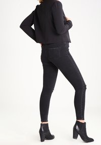 Femme portant un jean noir ajusté légèrement usé aux genoux, des bottines à talons noires et un top noir à manches longues court, debout de profil.