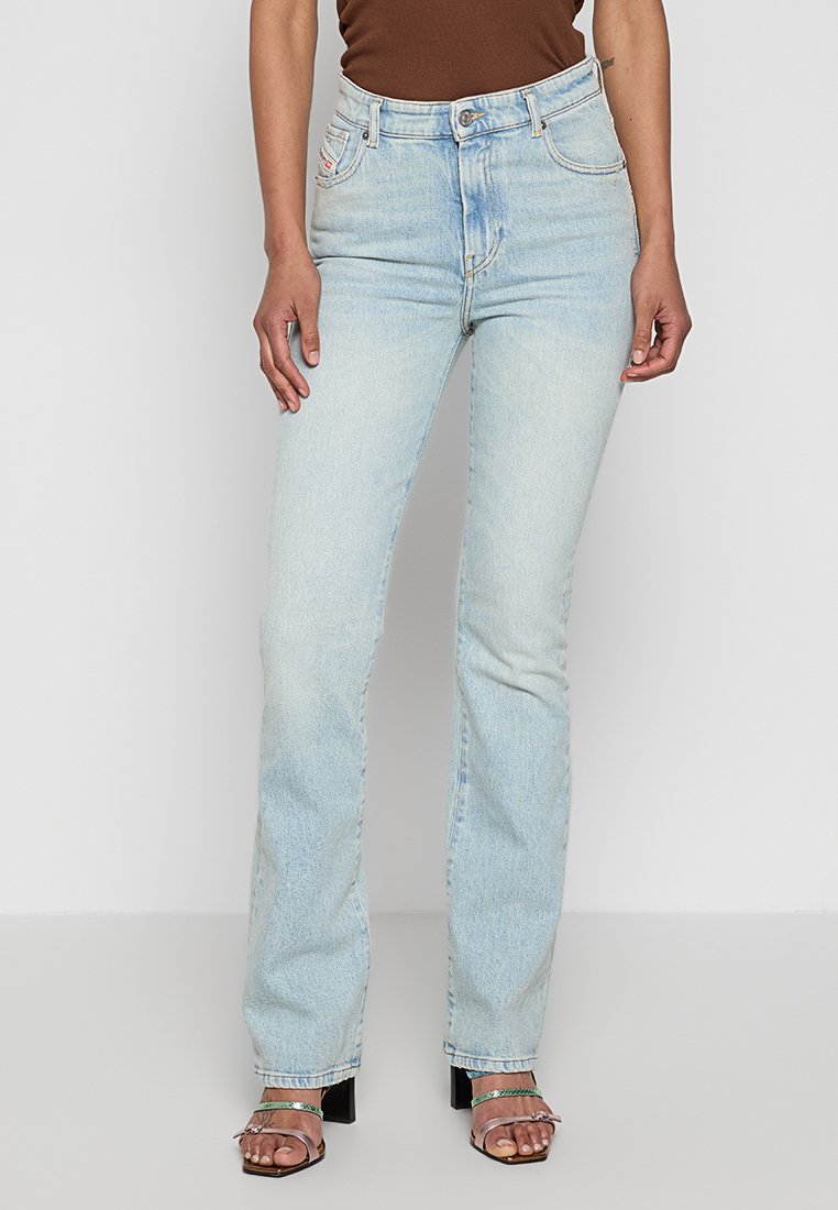 Helle blaue Jeans mit ausgestelltem Bein, fünf Taschen, Knopfverschluss und einer leicht abgenutzten Textur, kombiniert mit offenen Blockabsatzsandalen.