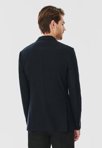 Granatowa marynarka o dopasowanym kroju, wykonana z teksturowanej tkaniny, z wyłogowym kołnierzem, pojedynczym rozcięciem z tyłu oraz zapinanymi mankietami, przedstawiona z widoku tyłu.