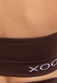 Banda de tejido acanalado marrón con una textura suave y detalle de logo "x" blanco, que exhibe un ajuste ceñido contra la piel.