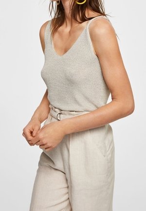 Kobieta ubrana w bezrękawnikowy beżowy dzianinowy top z dekoltem w serek oraz jasnobeżowe spodnie z wysokim stanem i materiałowym paskiem.