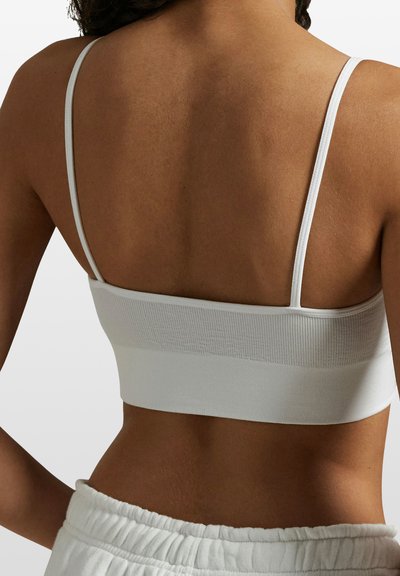 Bílá braletka s tenkými ramínky, žebrovanou texturou a širokým elastickým páskem na spodní části, se showcasing hladkým zadním designem.