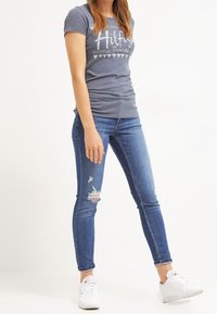 Camiseta gris con texto gráfico blanco, combinada con vaqueros ajustados rotos azules y zapatillas blancas. Atuendo casual.