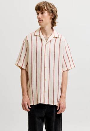 Jeune homme aux cheveux châtain clair portant une chemise blanche oversize à manches courtes avec des rayures rouges verticales et un pantalon noir, regardant sur le côté.