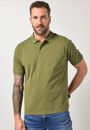 Homme aux cheveux bruns courts et barbe, portant un polo vert à motifs et un jean bleu, montrant son avant-bras gauche tatoué sur un fond clair.