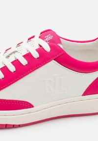 Baskets blanches avec des accents rose vif, des lacets blancs et un logo "RL" embossé sur le côté. Bord de semelle rose visible.