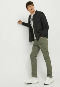 Camicia nera a maniche lunghe sopra una maglietta bianca con grafica, pantaloni verde oliva e scarpe da ginnastica bianche. Il modello è in piedi contro uno sfondo semplice.