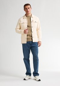 Chaqueta color crema con dos bolsillos en el pecho sobre un suéter marrón claro; unos vaqueros azules y zapatillas blancas completan el atuendo.