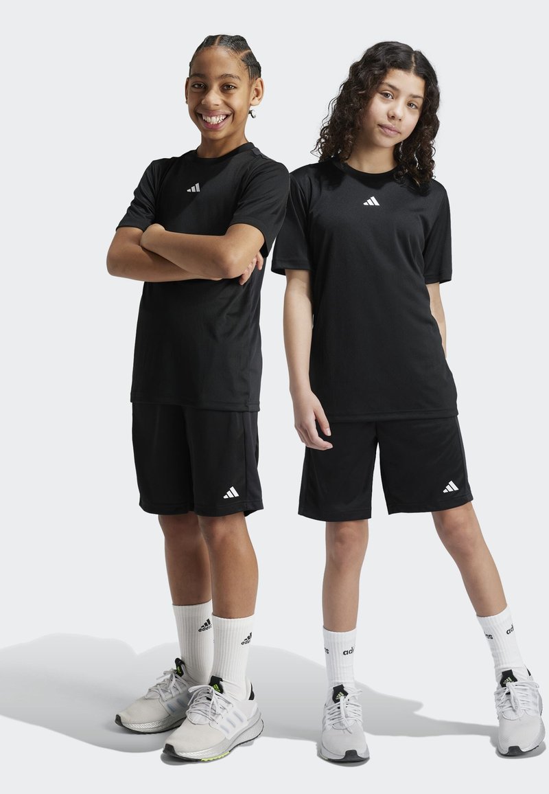 Dvaja chlapci majú na sebe zladené čierne športové oblečenie, vrátane tričiek a krátkych nohavíc, s bielymi akcentmi loga Adidas, doplnené bielymi ponožkami a športovými topánkami.