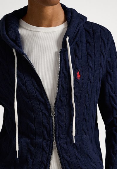 Temno modra kapa s kapuco z zadrgo in pletenim vzorcem, z belimi vrvicami, delno odprta čez belo rebrasto majico, na kateri je manjši rdeč vezen logotip.