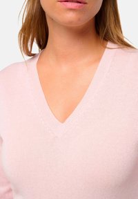 Gros plan d'une femme portant un pull en tricot rose clair à col en V, mettant en valeur le cou, les épaules et une partie de ses cheveux.