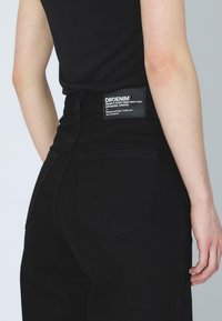 Vaqueros de mezclilla negros con cintura alta, con un bolsillo trasero rectangular y una etiqueta de marca, mostrados desde un lado y una vista trasera.