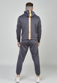 Completo con felpa grigia e pantaloni jogger, con una striscia d'accento dorata lungo la schiena, taglio slim, tessuto morbido e polsini a coste. Sneakers bianche completano il look.