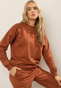 Blondynka w pomarańczowym swetrze i pasujących spodniach, siedząca z jedną ręką w kieszeni na beżowym tle.