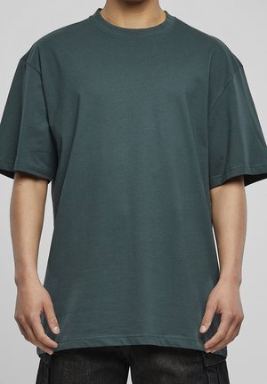 Mężczyzna w luźnym, ciemnozielonym t-shircie z krótkim rękawem i czarnych spodniach, stojący na jednolitym jasnoszarym tle.