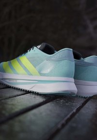 Jasnoniebieskie buty do biegania z neonowymi żółtymi paskami, siateczkową cholewką, szarymi akcentami i białą podeszwą środkową, wyeksponowane na ciemnej drewnianej powierzchni.