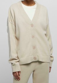 Beige stickad cardigan med v-ringning, fem knappar, ribbad krage och ärmslut. Mjukt material, något oversized passform, tillsammans med en ljus topp.